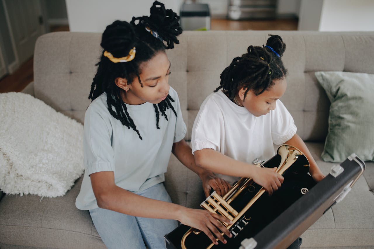 Home Two young girls with dreadlocks exploring a trumpet together indoors, fostering music interest.